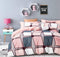 A bedroom scene with a bed having a multicolored geometric patterned top sheet. The room is decorated with pink accents, a nightstand, a lamp, and framed pictures on the wall.